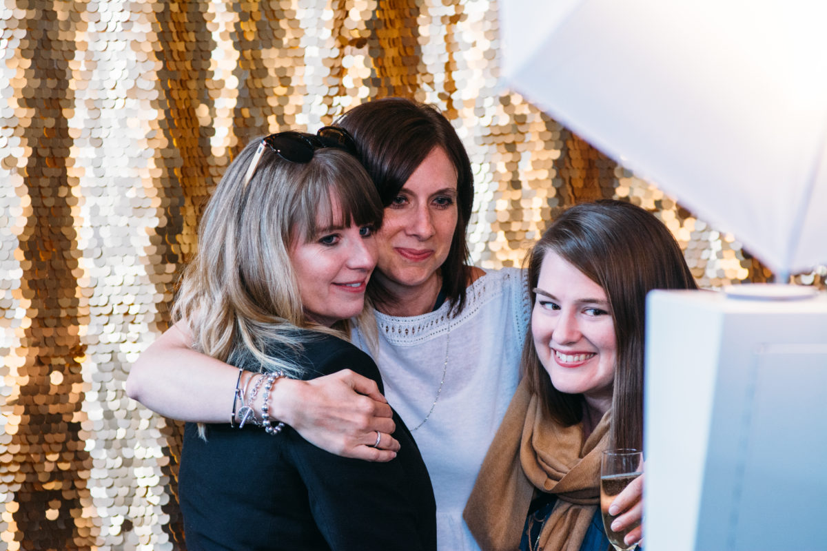 Three Unico employees gather in front of a shiny gold backdrop to pose for a photo booth snapshot