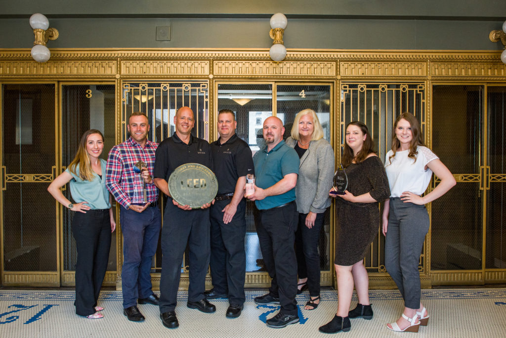 Property team at Smith Tower in front of the building's iconic historic elevators with the building's various awards including: LEED Platinum plaque, local TOBY Award, regional TOBY Award, and international TOBY Award. Smith Tower was acquired by real estate investment company Unico in 2015.